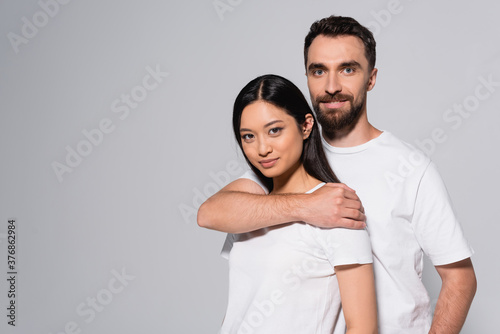 bearded man in white t-shirt embracing young asian woman while posing isolated on grey