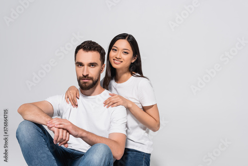 brunette asian woman touching boyfriend and looking at camera while sitting together isolated on white