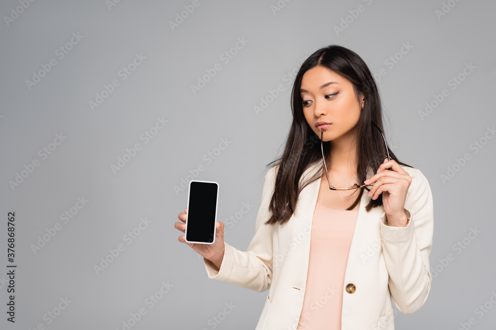 thoughtful asian businesswoman in blazer holding eyeglasses while showing smartphone with blank screen isolated on grey thoughtful asian businesswoman in blazer holding eyeglasses while showing smartphone with blank screen isolated on grey