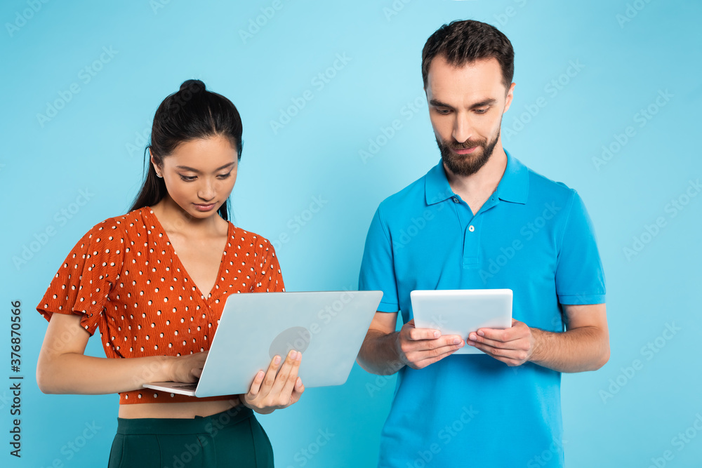 young asian woman in red blouse using laptop near bearded man with digital tablet on blue young asian woman in red blouse using laptop near bearded man with digital tablet on blue