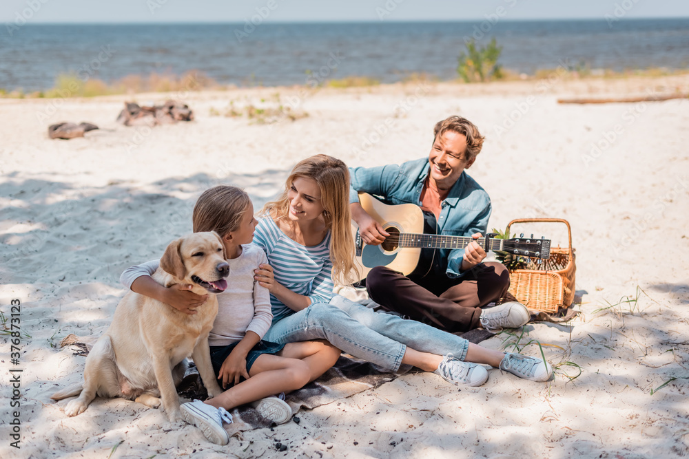 Woman hugging daughter with golden retriever while husband playing acoustic guitar on beach Woman hugging daughter with golden retriever while husband playing acoustic guitar on beach