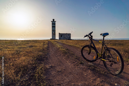 The bike stands on the road in front of the old lighthouse on the seashore, in the rays of the setting sun.
