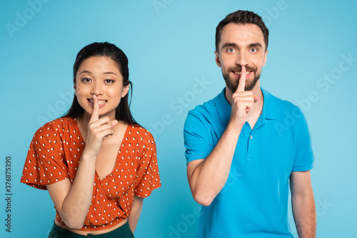young interracial couple looking at camera while showing hush gesture on blue