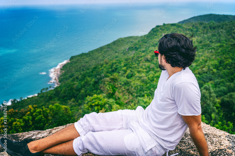 Young handsome hiker in white t-shirt and shorts admires the beautiful ocean view from the top of the mountain. Vacation travel concept