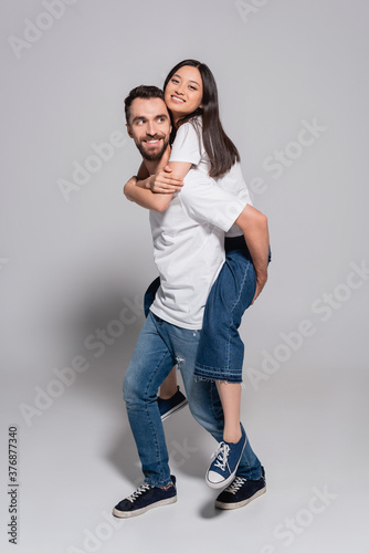 bearded man in white t-shirt and jeans piggybacking asian girlfriend on grey