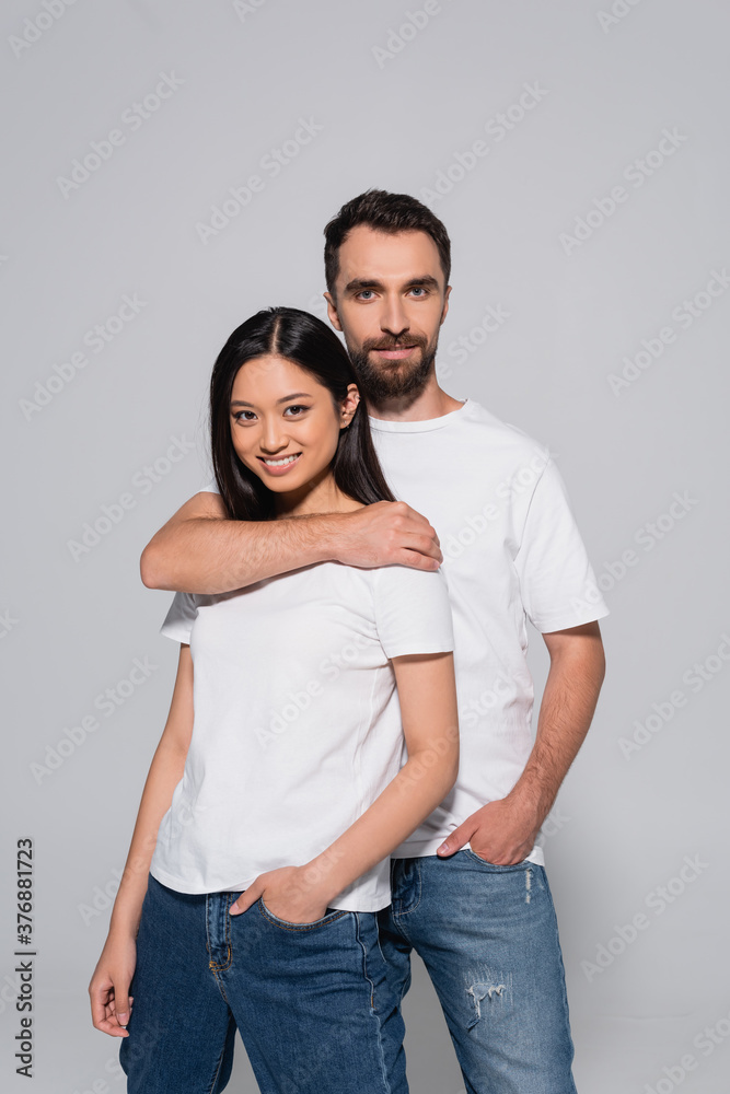 bearded man hugging brunette asian woman while posing with hands in pockets isolated on grey bearded man hugging brunette asian woman while posing with hands in pockets isolated on grey