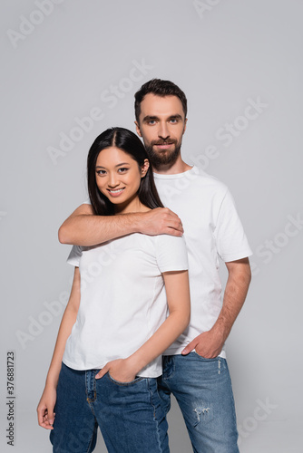 bearded man hugging brunette asian woman while posing with hands in pockets isolated on grey