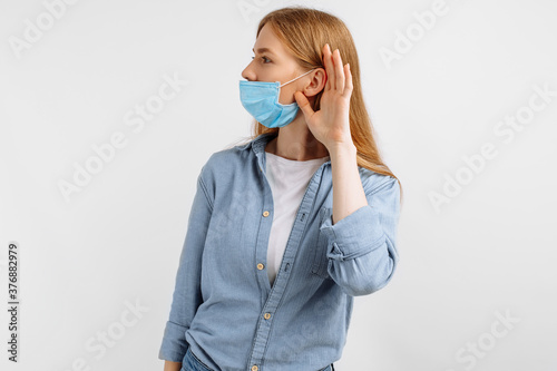 curious, focused young woman in a medical protective mask on her face, listening with her hand to her ear, against a white background
