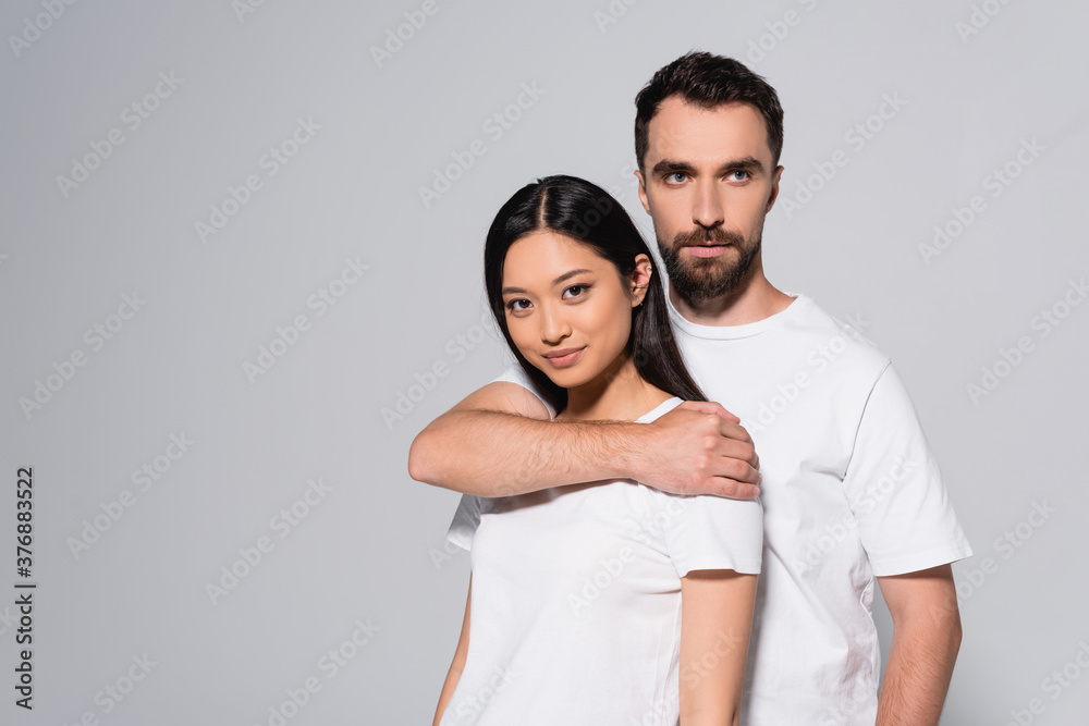 young man in white t-shirt hugging asian woman while posing isolated on grey young man in white t-shirt hugging asian woman while posing isolated on grey