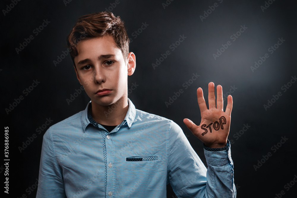 The concept of domestic violence. A sad teenage boy shows a palm up that says STOP. Copy space. Black background