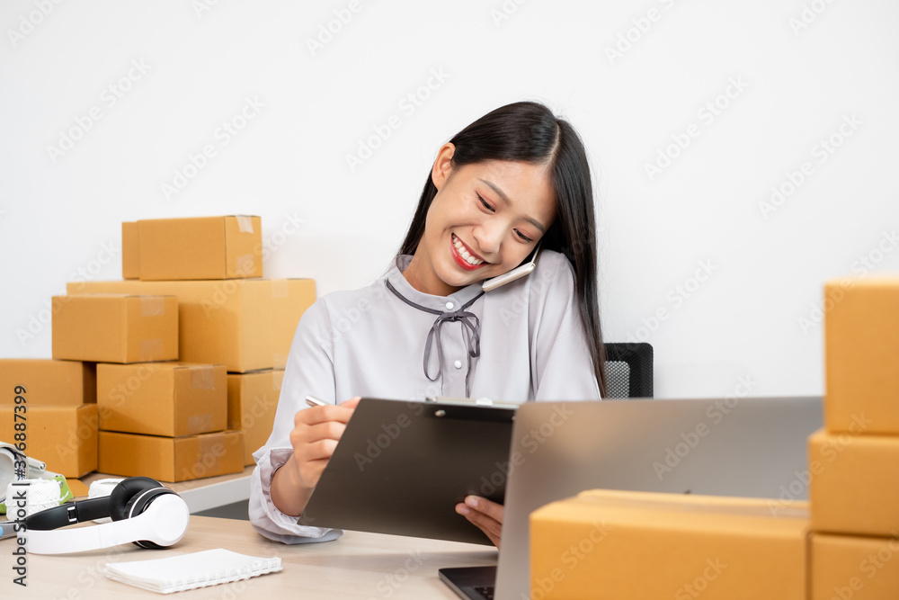 Young Asian woman business owner with many parcel boxes on the table happy online sales job, use your smartphone, get an order from customers, take notes, and make arrangements for delivery by post Young Asian woman business owner with many parcel boxes on the table happy online sales job, use your smartphone, get an order from customers, take notes, and make arrangements for delivery by post