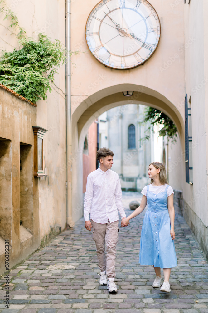 Outdoor city portrait of happy couple in love, handsome boyfriend and attractive young girl in blue dress, walking together in beautiful European city, holding hands and smiling