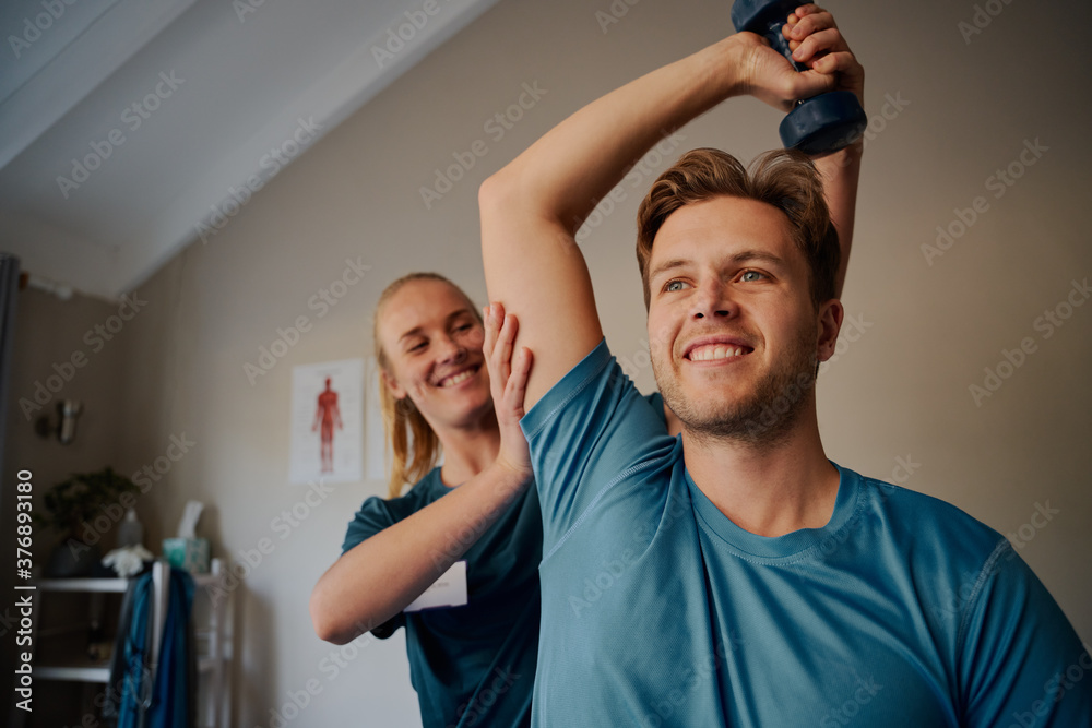 Obraz premium Portrait of handsome young smiling man exercising using dumbbells with female physiotherapist 