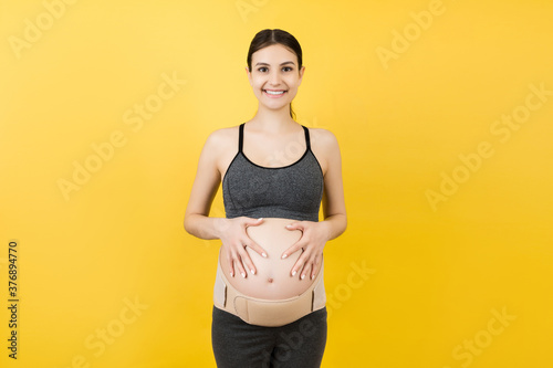 Close up of pregnant woman in underwear wearing elastic pregnancy bandage to make the pain go away at yellow background with copy space. Orthopedic abdominal support belt concept © sosiukin
