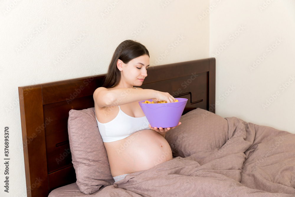 Pregnant woman relaxing in bed is eating chips because of salt cravings. Unhealthy junk food during pregnancy concept Pregnant woman relaxing in bed is eating chips because of salt cravings. Unhealthy junk food during pregnancy concept