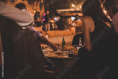 Persone di spalle sedute in un locale serale all'aperto degustano cibo e bevande su tavolo di legno photo