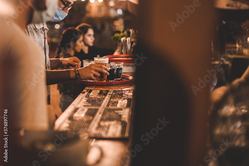 Barista con mascherina di sera serve su vassoio una birra al bancone di un bar in legno all'aperto photo