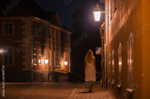 One young alone woman in white dress slowly walking on sidewalk under street lights at old town in black summer night. Peaceful atmosphere. Spending time alone. City life. Back view.