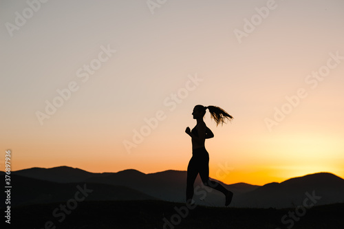 Silhouette against sky at sunrise while running. Runner fitness girl in sport tight clothes. Athlete woman on the road in the mountains. Bright sunset and blurry background. Workout wellness concept.