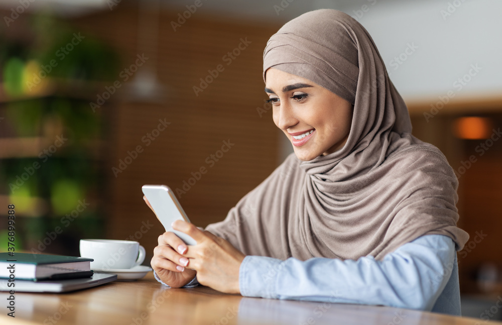 Smiling girl in hijab using smartphone while resting at cafe Smiling girl in hijab using smartphone while resting at cafe