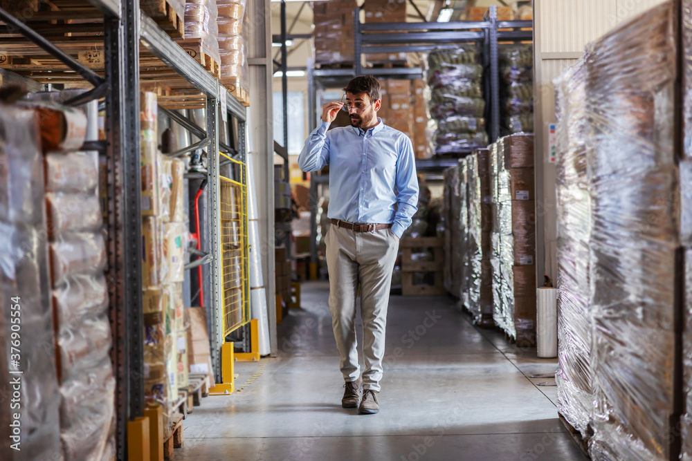 Full length of young attractive CEO walking trough warehouse and checking on goods.