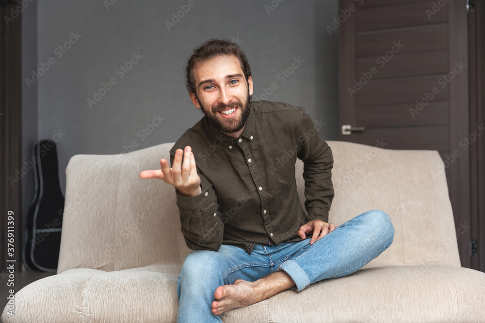 Positive handsome man with a beard talks and gestures while sitting on the couch. The guy looks at the camera