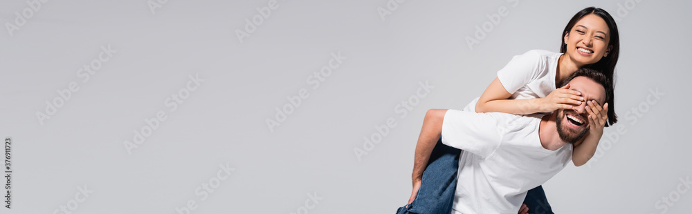 horizontal image of laughing man piggybacking asian girlfriend covering his eyes with hands isolated on grey horizontal image of laughing man piggybacking asian girlfriend covering his eyes with hands isolated on grey