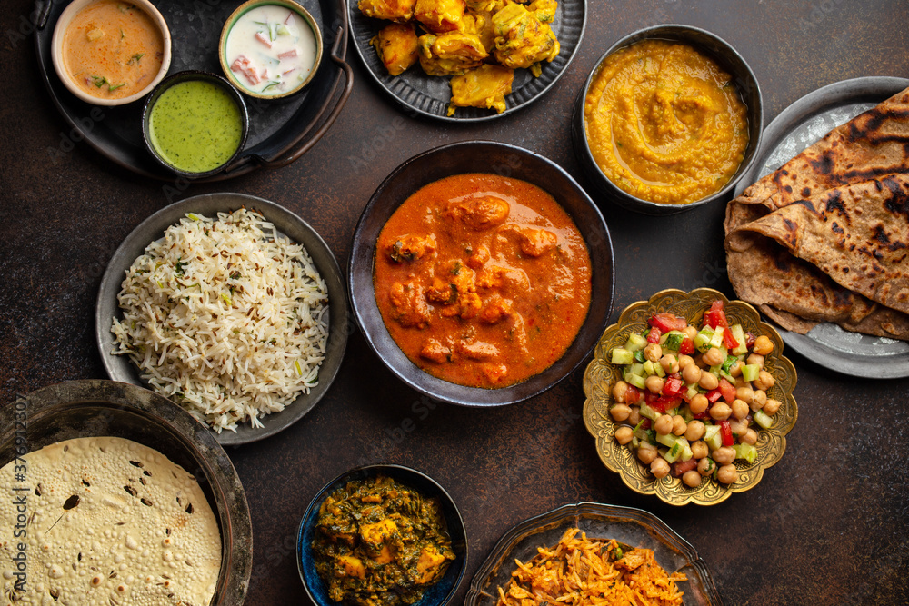 Variety of Indian food, different dishes and snacks on dark rustic background. Pilaf, butter chicken curry, rice, palak paneer, chicken tikka, dal soup, naan bread, assortment of chutney. Top view.