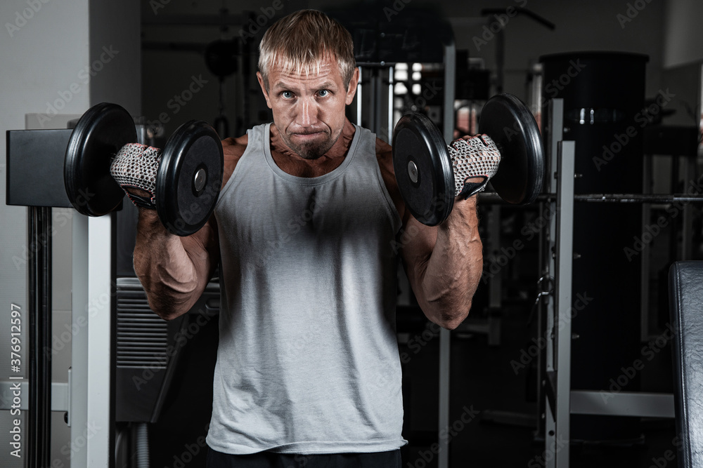 Muscular adult brutal man with dumbbells pumps biceps in the gym. Portrait of caucasian authentic bodybuilder doing workout exercises Muscular adult brutal man with dumbbells pumps biceps in the gym. Portrait of caucasian authentic bodybuilder doing workout exercises