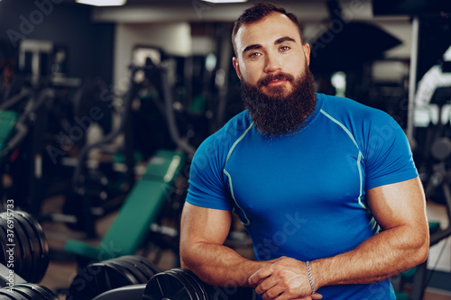 Bearded young man bodybuilder in blue t-shirt standing in gym