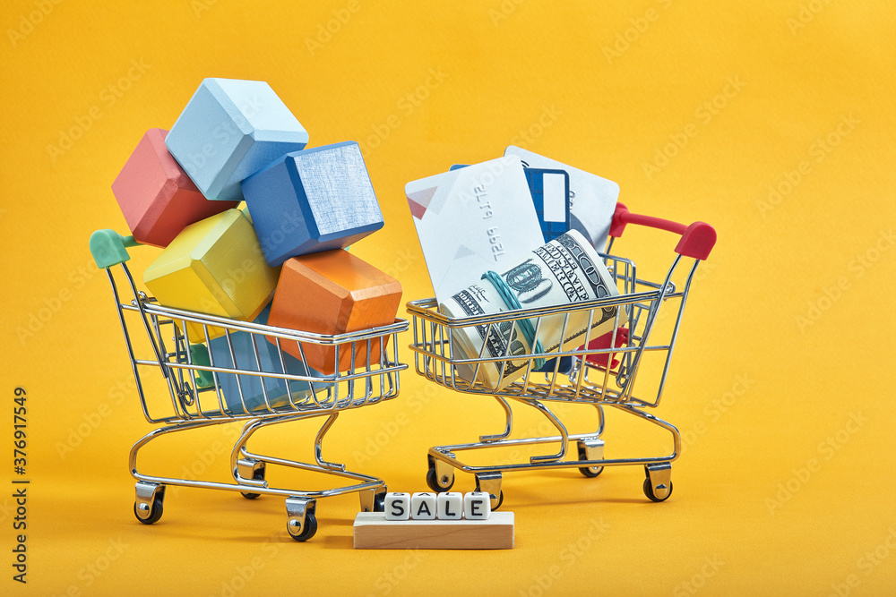 two shopping trolleys with multi-colored cubes, credit cards and dollars on yellow background. Sales word written on cubes.