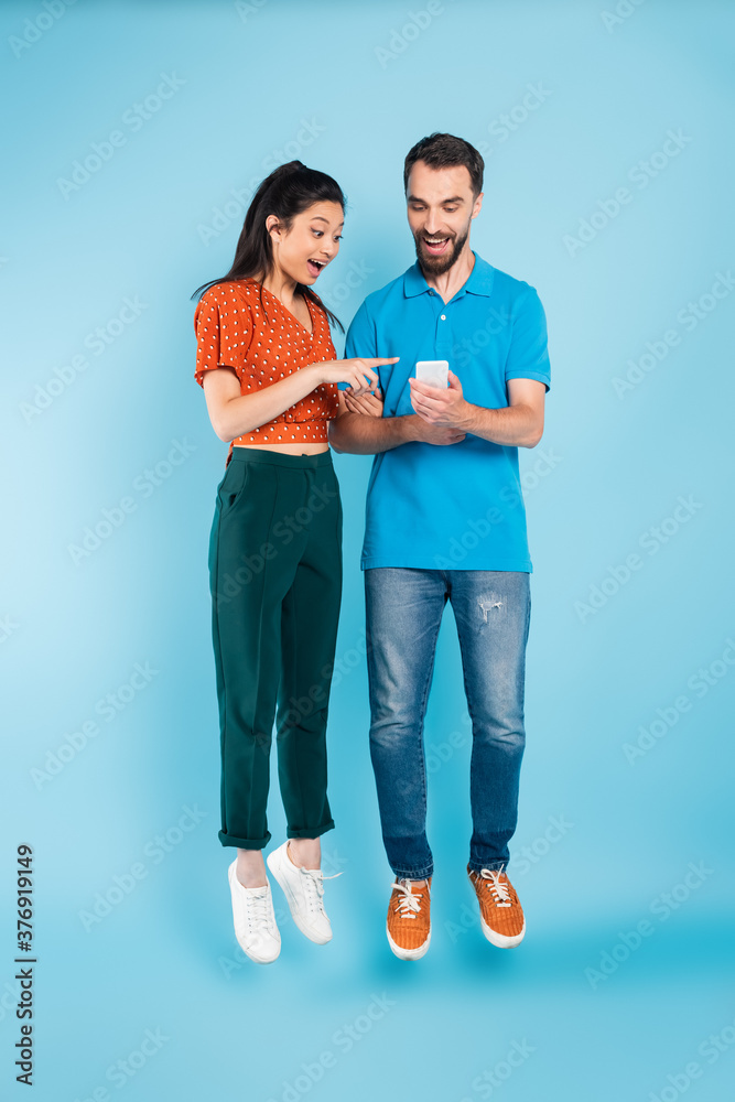 excited asian woman pointing with finger at smartphone in hands of bearded man while levitating on blue