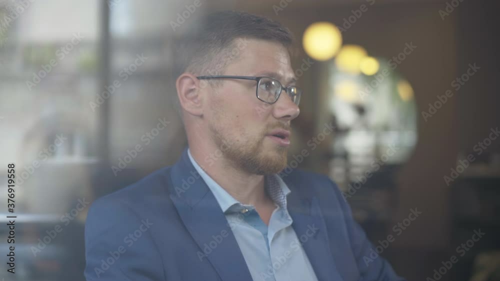 Close-up of confident Caucasian businessman in eyeglasses talking to unknown person. Portrait of young handsome man sitting in cafe behind glass window and chatting. Business lifestyle. Close-up of confident Caucasian businessman in eyeglasses talking to unknown person. Portrait of young handsome man sitting in cafe behind glass window and chatting. Business lifestyle.