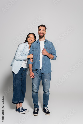 full length view of stylish asian woman leaning on bearded boyfriend in denim clothes on grey