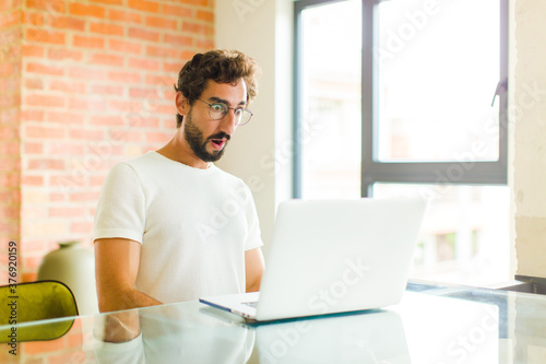 young bearded man with a laptop looking very shocked or surprised, staring with open mouth saying wow photo