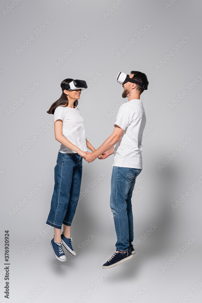 young couple in jeans and white t-shirts holding hands while levitating in vr headsets on grey
