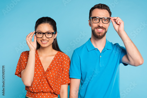young interracial couple looking at camera while touching eyeglasses isolated on blue