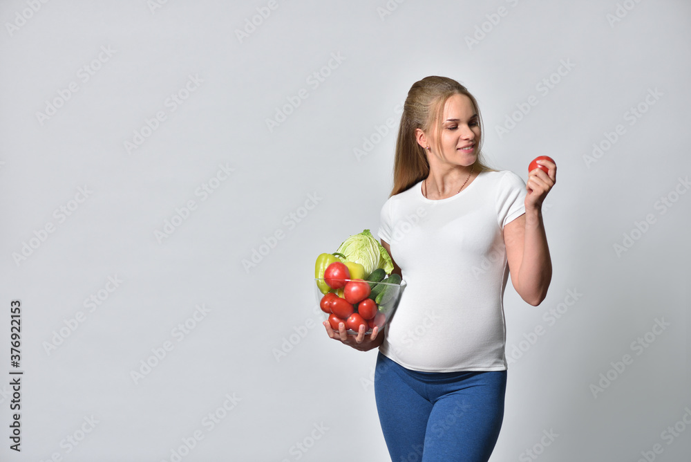 Pregnant blonde woman holding fresh vegetables in a bowl and tomato in her hand Pregnant blonde woman holding fresh vegetables in a bowl and tomato in her hand