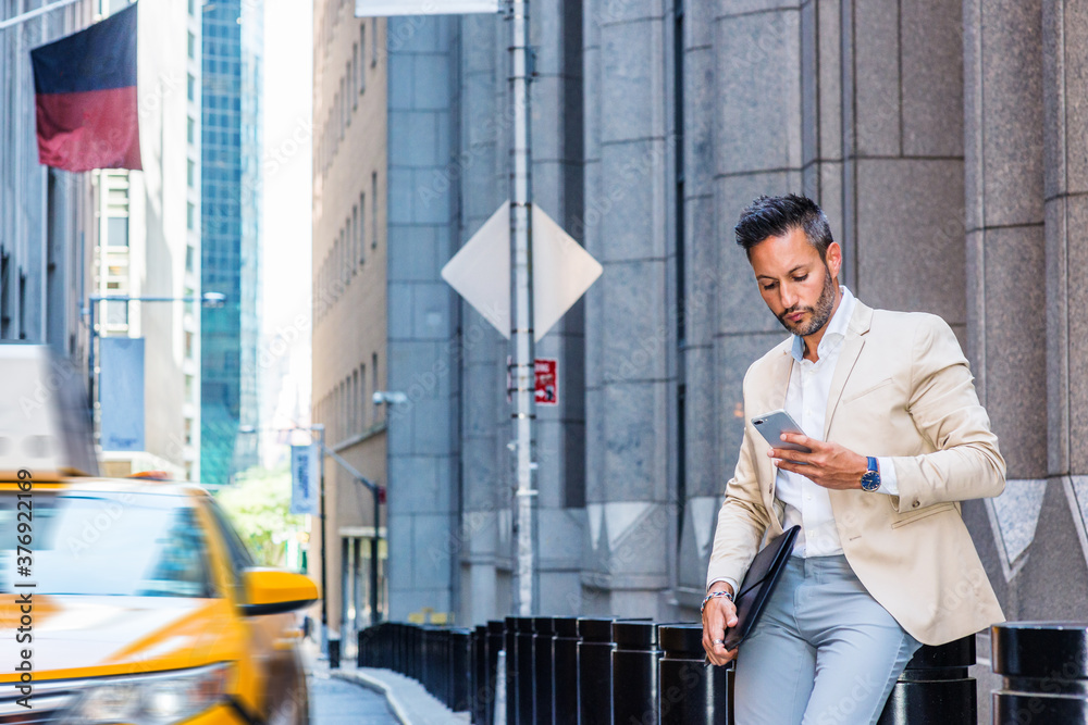 Young European Businessman traveling, working in New York, with beard, little gray hair, wearing beige blazer, gray pants, holding briefcase, sitting on old street, looking down, texting on cell phone