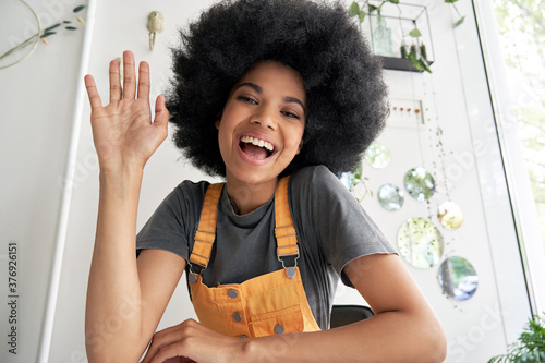 Pretty young African American mixed race hipster vlogger woman with Afro hair waving hand looking at webcam talking to camera sits at cafe table video calling, recording blog, headshot portrait. photo