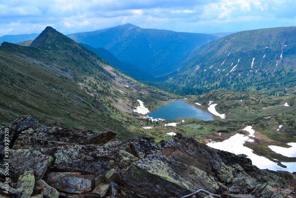 stones, view of mountain lake in form of heart among snow ice glaciers, green mountain ranges overgrown with trees, Chersky peak