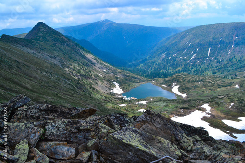 stones, view of mountain lake in form of heart among snow ice glaciers, green mountain ranges overgrown with trees, Chersky peak