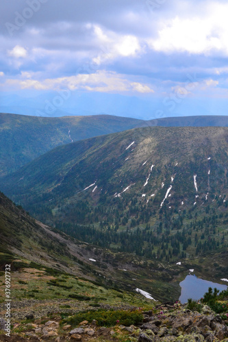 view of mountain blue lake among snowy green mountain ranges overgrown with trees, Chersky peak, clouds