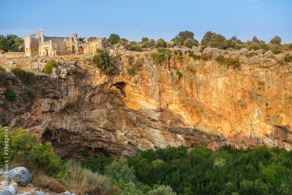 Panorama of ruins, lighted by sun, in ancient city Canytelis or Kanlıdivane, Ayaş, Turkey. Church built in 2 century AD near edge of karst sinkhole. Diameter of hole about 130 m, depth around 70 m.