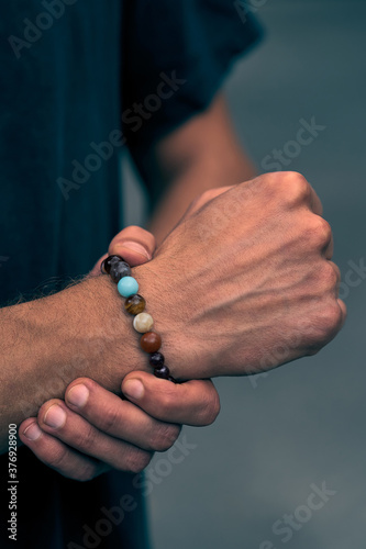 A man in dark blue t-shirt put his fingers around wrist with stylish beaded bracelet. The beads are carved of colorful ornamental stones. Cropped shot on the gray background. 