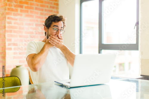 young bearded man with a laptop covering mouth with hands with a shocked, surprised expression, keeping a secret or saying oops