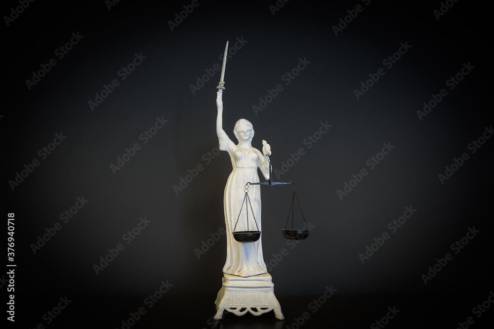 White figurine on black background of the goddess of a Themis with scales in a hand and a bandage on eyes. Justice Scales Law Lawyer Business Concept. White figurine on black background of the goddess of a Themis with scales in a hand and a bandage on eyes. Justice Scales Law Lawyer Business Concept.