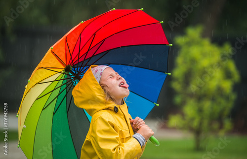 happy kid catching rain drops in park. Funny little girl playing under the autumn rain. Kid wearing yellow waterproof coat and boots holding colorful umbrella.