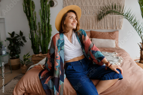 Fascinating brunette  woman in straw hat chilling at home in cozy boho interior. Macrame on wall. Palm trees and cactus on background.