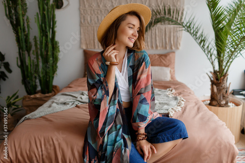 Smiling  woman in straw hat chilling at home in cozy boho interior. Macrame on wall. Palm trees and cactus on background.